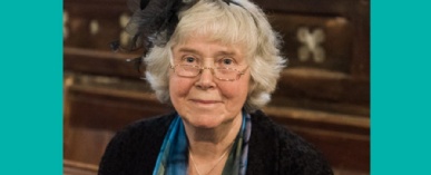 A grey-haired woman dressed in blue for a wedding and wearing glasses, smiles at the camera from her pew.