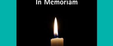 Close up image of the top of a lit candle with the flame shown against a dark background and the words In Memoriam written above in white text.